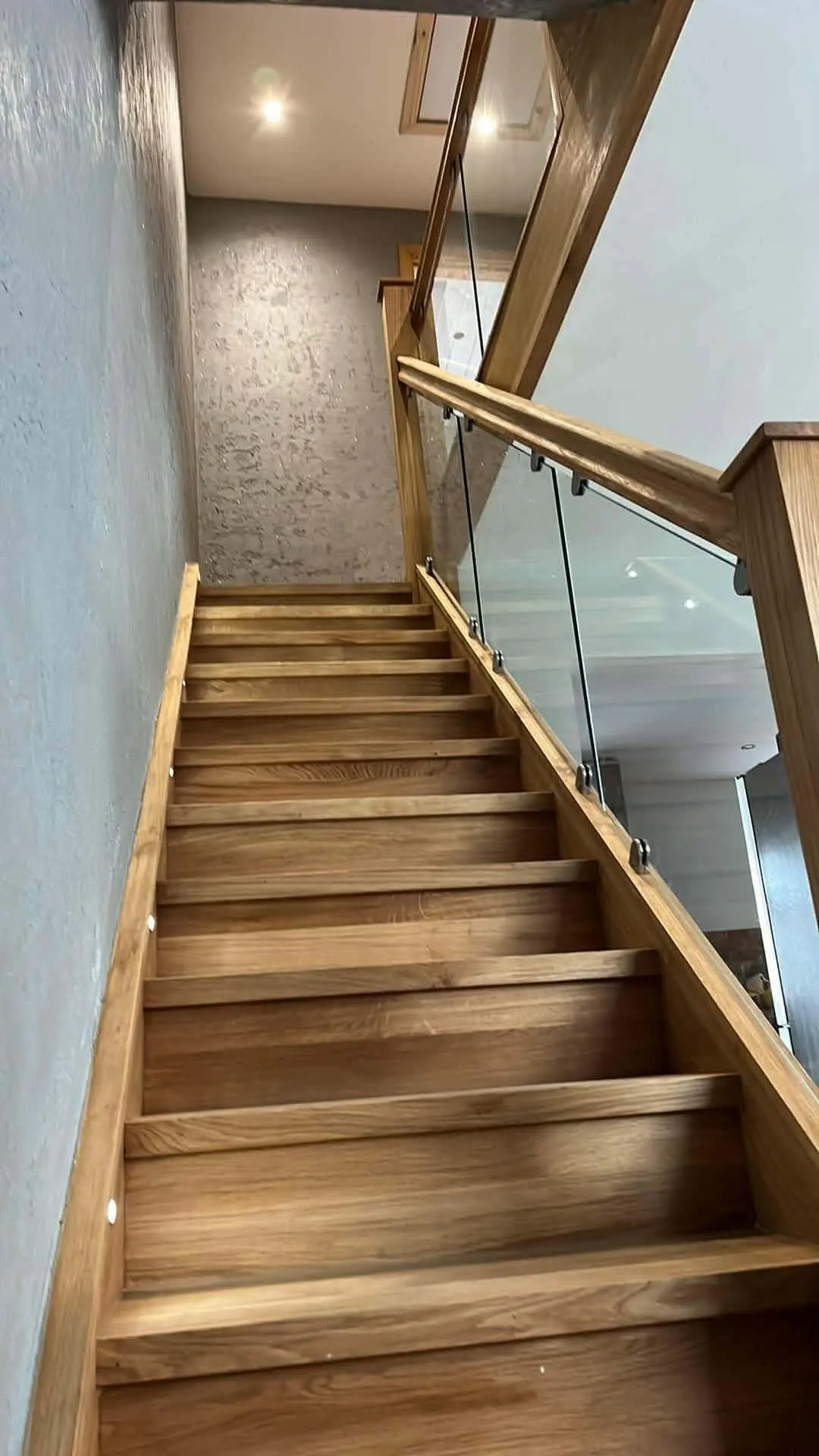 Long timber staircase with a glass balustrade and timber handrail, seen from the foot of the stairs